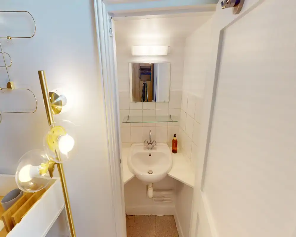 Petit coin lavabo de salle de bains avec vasque ronde, miroir mural, étagère en verre et éclairage chaud — propre et bien éclairé.