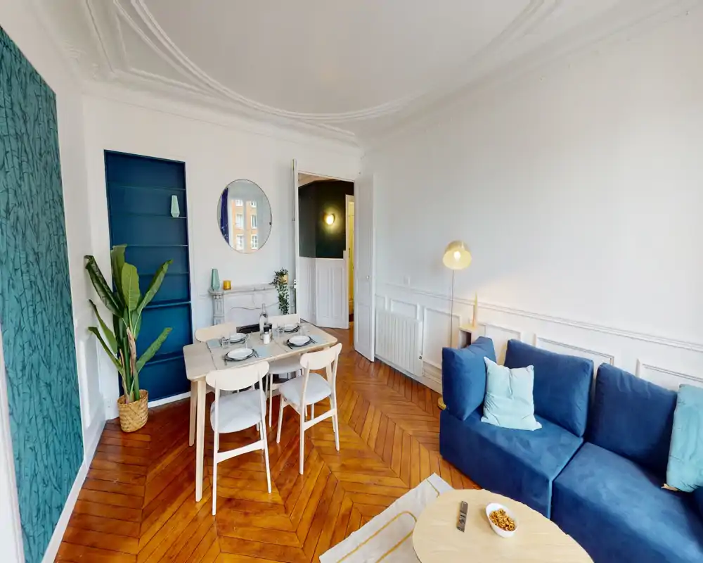 Salon lumineux et élégant avec un canapé bleu, parquet en bois et une petite table à manger dressée pour quatre. L'espace présente moulures décoratives, une plante et des accents soignés pour une ambiance chaleureuse et moderne.