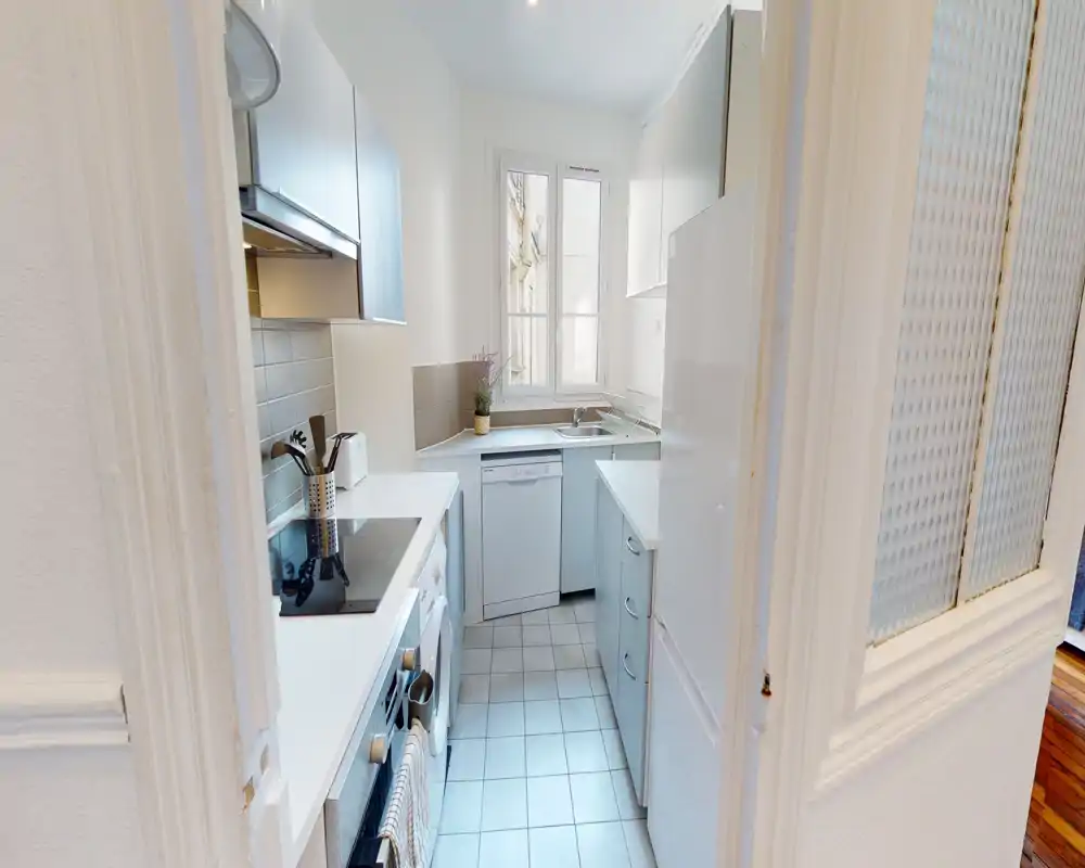 Cuisine en enfilade compacte et moderne avec placards blancs, électroménagers intégrés et une fenêtre apportant de la lumière naturelle ; plans de travail rangés et petite plante conviviale.