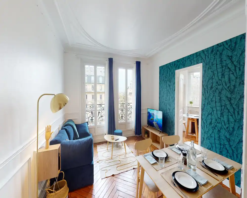 Salon lumineux et stylé avec canapé bleu marine, mur d'accent texturé, table à manger dressée pour quatre et grandes fenêtres donnant sur un balcon classique — clair, soigné et chaleureux.