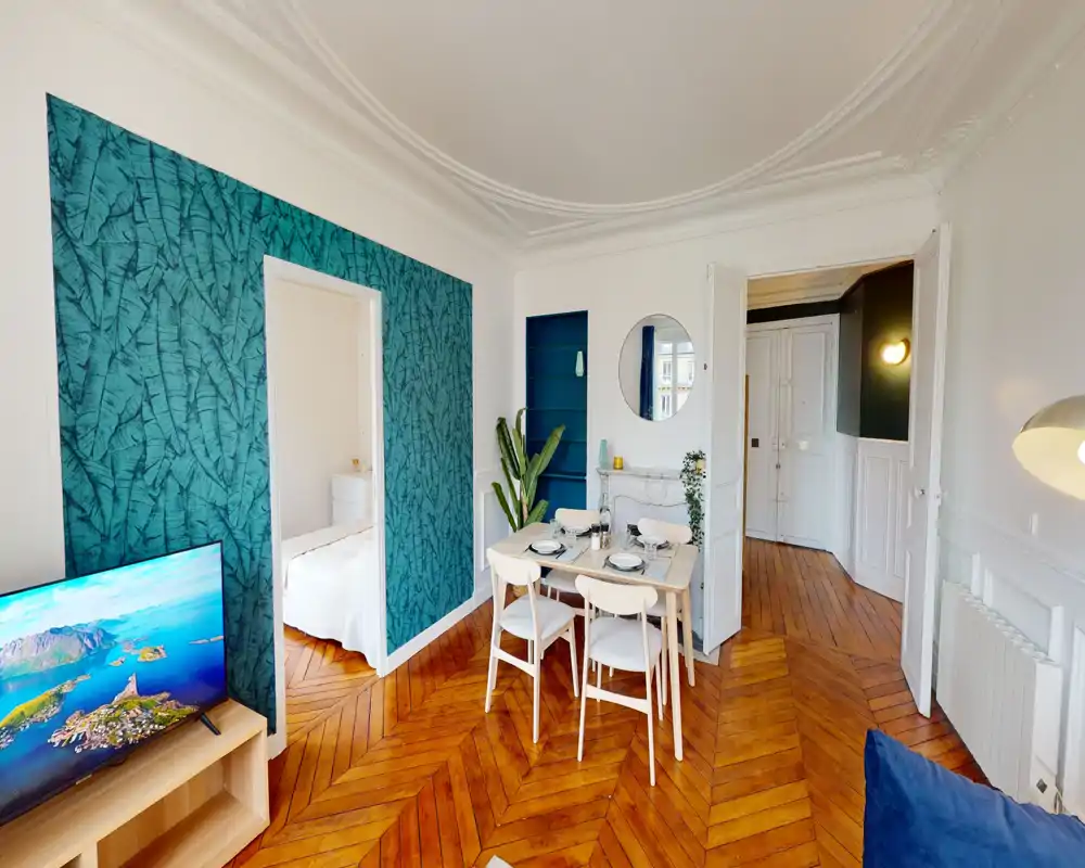Pièce à vivre lumineuse et élégante avec petite table à manger, télévision et mur d'accent teal. Parquet en chevrons et décoration soignée créent une ambiance chaleureuse et accueillante, idéale pour une annonce immobilière.
