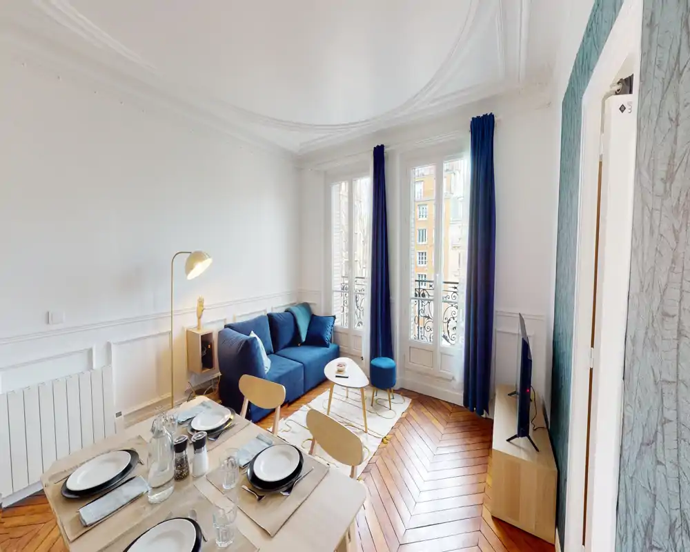 Séjour lumineux et élégant avec canapé bleu, lumière naturelle provenant de grandes fenêtres à la française et petite zone repas au premier plan.
