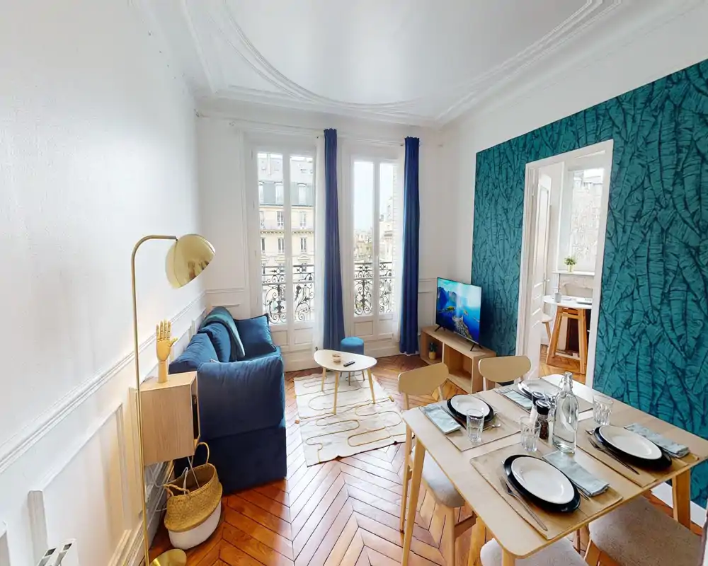 Salon lumineux et élégant avec canapé bleu, mur d'accent à motif et table à manger pour quatre ; grandes fenêtres avec balcon apportant une belle lumière naturelle.