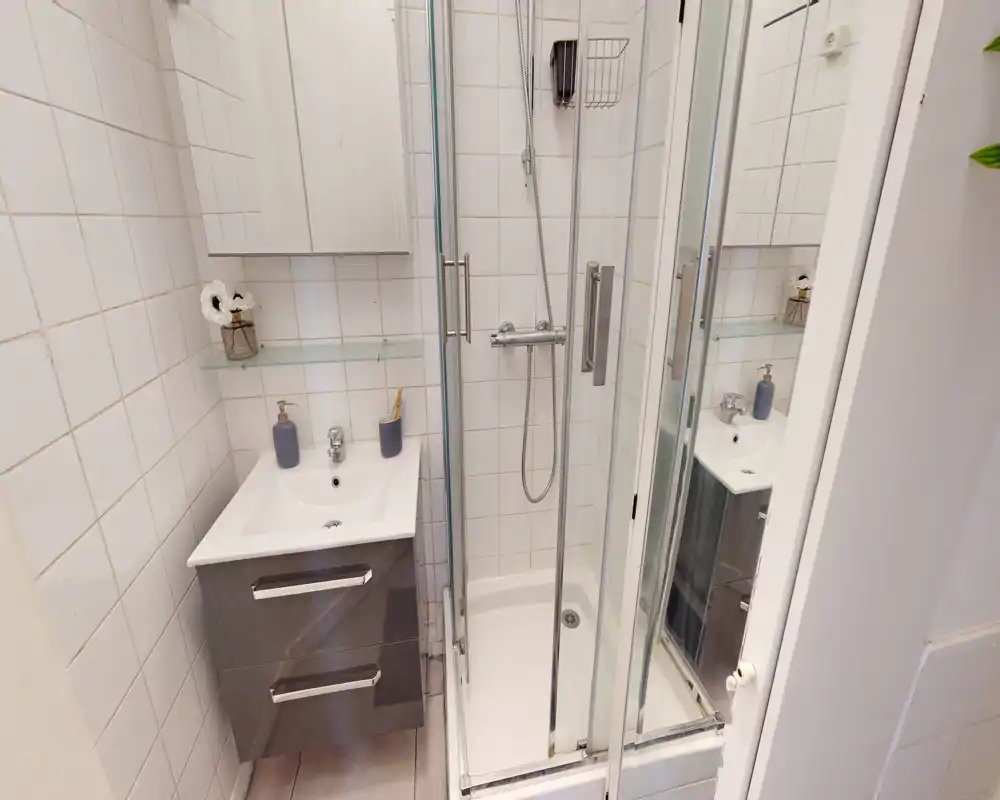 Salle de bains compacte avec douche vitrée, meuble lavabo mural et armoire miroir — carrelage blanc et équipements modernes.