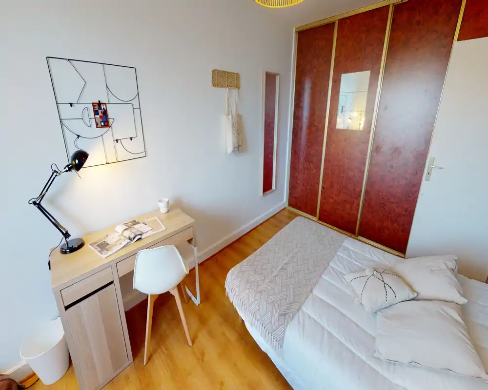 Dormitorio luminoso con cama doble bien hecha, manta y cojines, y un escritorio compacto de madera junto a la pared: un espacio acogedor y funcional para una o dos personas.