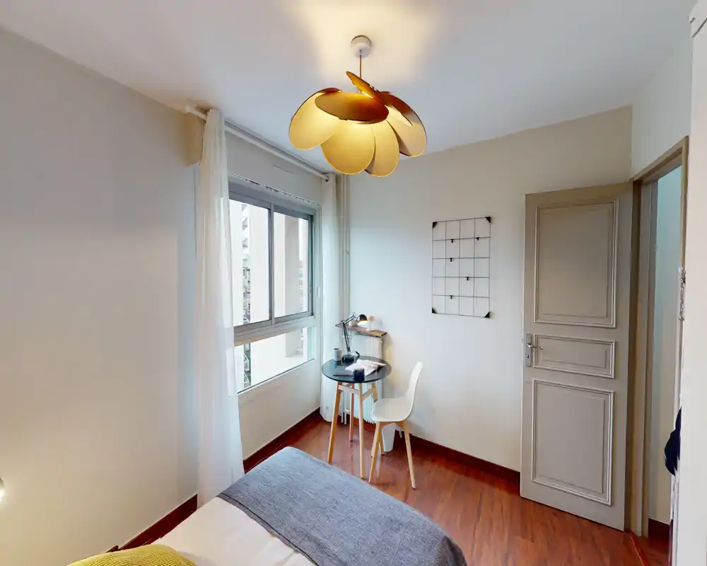 Chambre simple et chaleureuse avec lit visible, parquet et petit bureau près de la fenêtre. Luminaire moderne et voilages apportent une ambiance lumineuse et accueillante, idéale pour les locations courte durée.