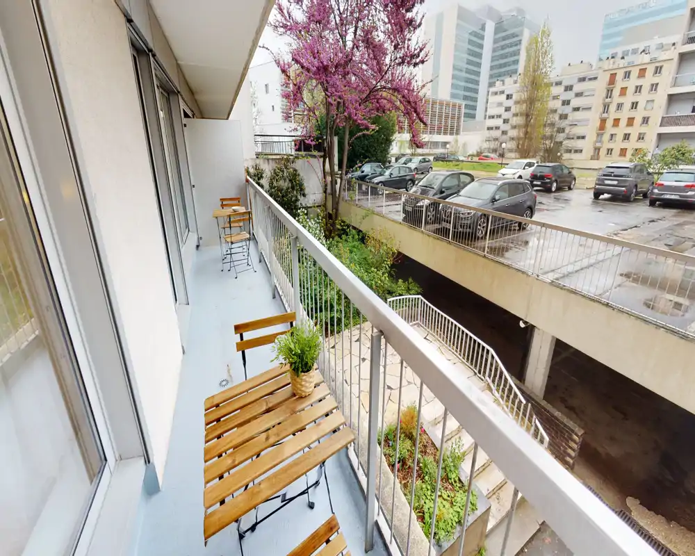 Balcone con piccolo tavolo e sedie in legno, pianta in vaso e ringhiera che si affaccia su un cortile residenziale e parcheggio — spazio esterno compatto e sfruttabile.