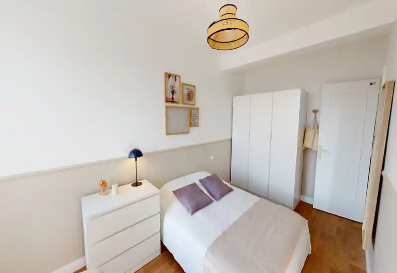 Bright, neatly styled bedroom with a double bed, bedside lamp, dresser and wardrobe; warm neutral tones and soft textiles create a calm atmosphere.