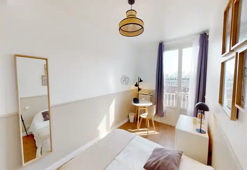 Bright, cozy bedroom with a comfortable double bed, soft linens, a bedside chest with lamp, a small desk by the window and a large mirror — ideal for a single occupant or couple seeking a sunny room.