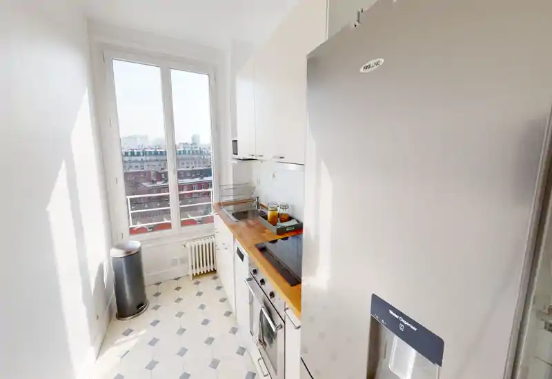 Cuisine en enfilade lumineuse et compacte avec plan de travail en bois, plaque à induction, réfrigérateur inox et grande fenêtre offrant une vue sur la ville.