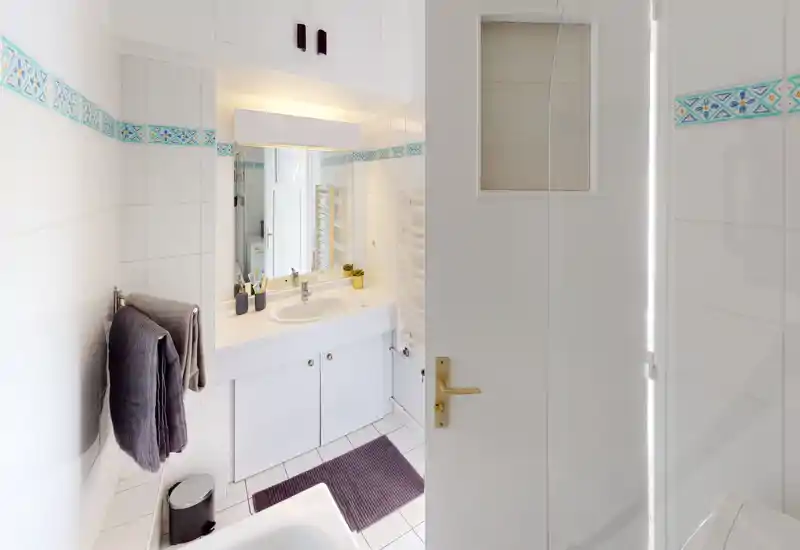 Bright, clean bathroom with a sink, mirror, towel rack and tiled walls. Well-lit white tiles and decorative border tiles give a fresh look.