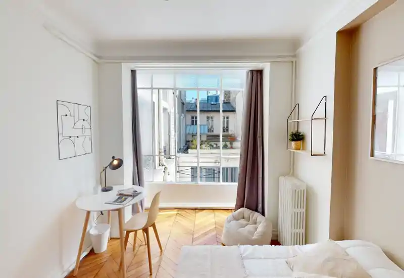Bright, stylish bedroom with a comfortable bed in the foreground, a small workspace by the window and warm wooden chevron floors. Large windows provide plenty of natural light and a pleasant view of neighboring rooftop planters.