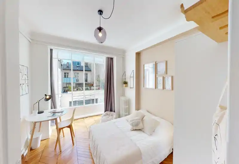 Bright, cozy bedroom with a comfortable double bed, soft textiles and natural light from a large window; simple desk area by the window and warm wooden parquet flooring.