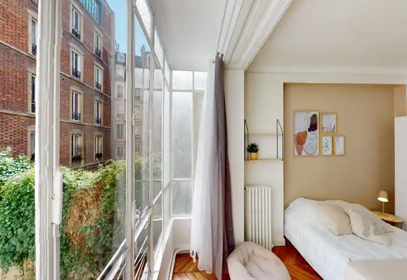 Bright, cozy bedroom with a single bed beside a large French-window that fills the room with natural light; warm wooden floor and soft textiles create a welcoming atmosphere.