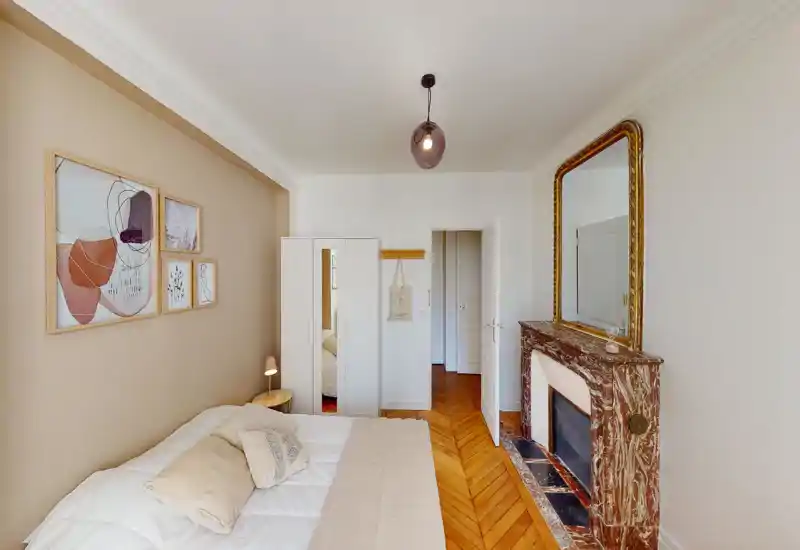 Bright, stylish bedroom with a comfortable double bed, soft neutral textiles and a decorative marble fireplace with large mirror. Warm wooden parquet and framed wall art complete the cozy, elegant atmosphere.