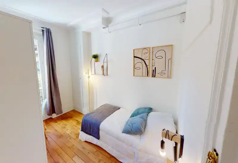 Bright, compact bedroom with a single bed dressed in white linen, blue cushions and a grey throw. The room features warm wooden floors, a floor lamp, simple wall art and a window with curtains letting in natural light.