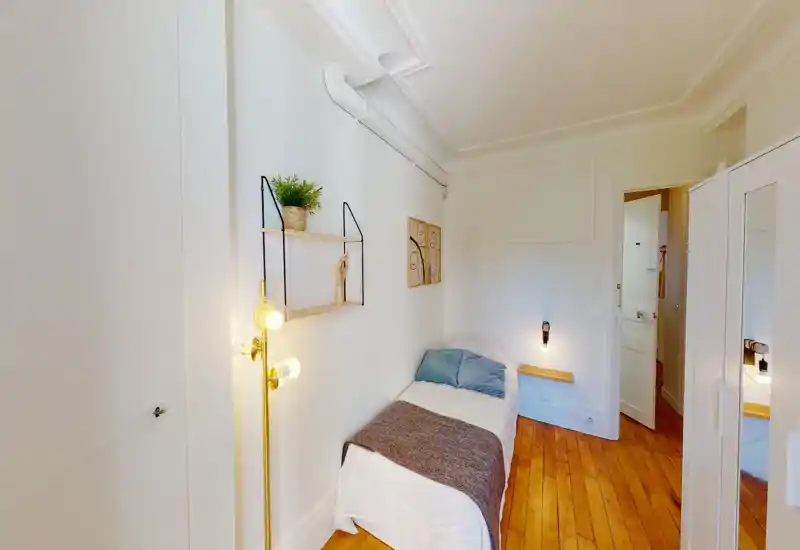 Bright, compact single bedroom with a neatly made bed, soft pillows and a textured throw. Warm wooden floors, tasteful wall shelving and ambient lighting create a cozy, well-styled space ideal for a single occupant.