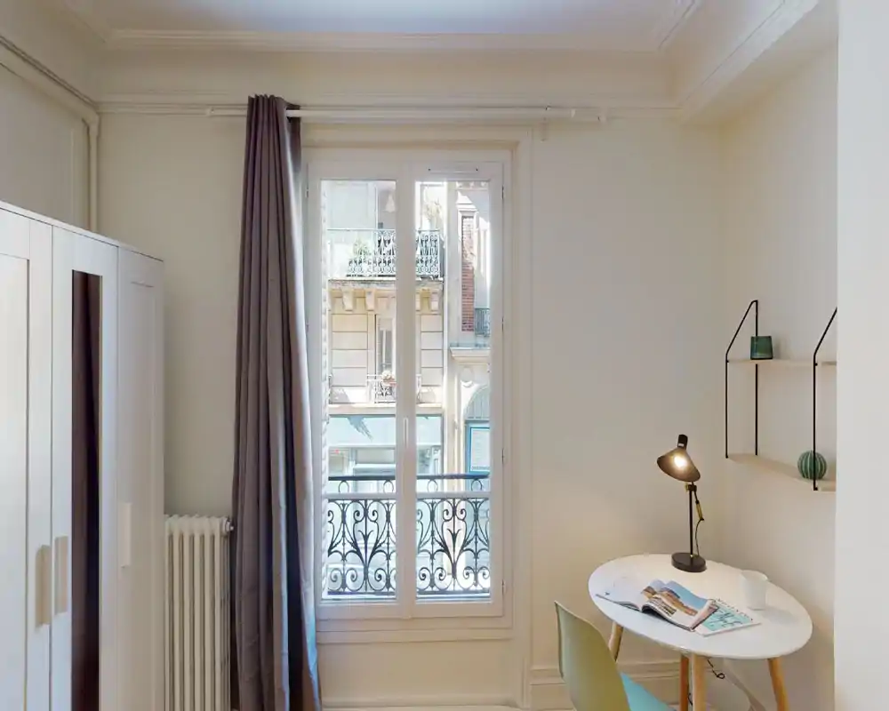 Bright, minimal bedroom corner with a large window and small work table. Neutral walls, full-length curtains and a wardrobe create a calm, functional space ideal for a single occupant.