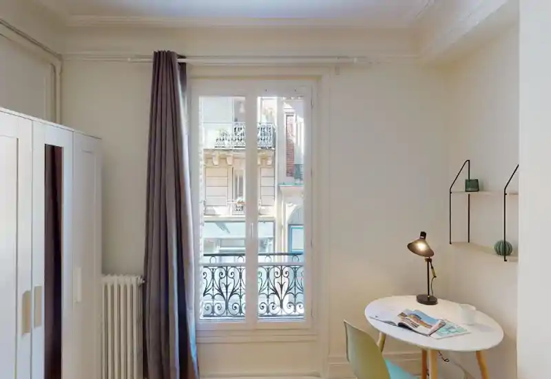 Bright, minimal bedroom corner with a large window and small work table. Neutral walls, full-length curtains and a wardrobe create a calm, functional space ideal for a single occupant.