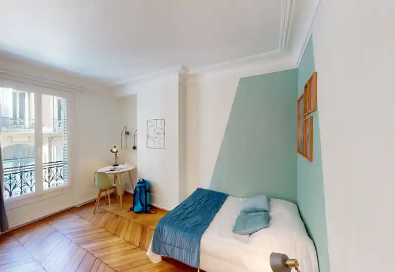 Sunny, stylish bedroom with a double bed against a mint-green accent wall, soft bedding and a small workspace by a large window providing abundant natural light.