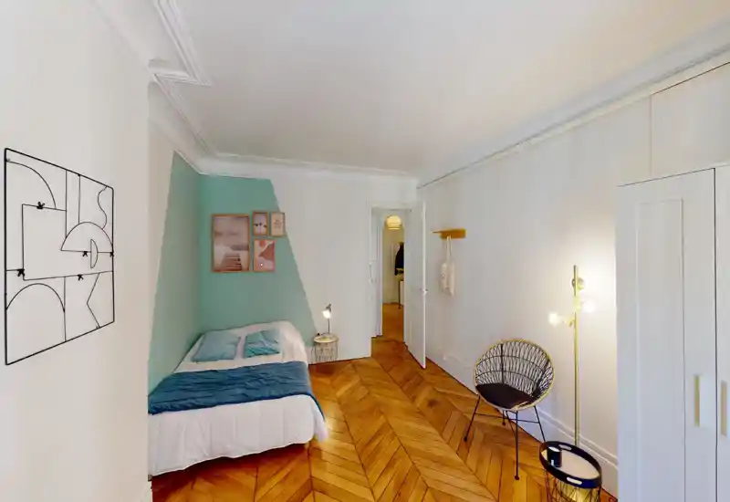 Bright, stylish bedroom with a double bed set against a mint-accented wall, warm chevron wooden floors and tasteful wall art. A cozy bedside lamp and a modern rattan chair complete the inviting space.