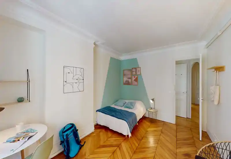 Bright, stylish bedroom with a double bed against a painted accent wall, coordinated teal bedding and framed artwork; hardwood herringbone floors and a bedside lamp complete the cozy setup.