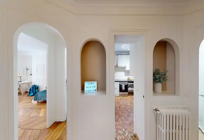 Bright hallway with arched openings showing a bedroom on the left and a kitchen straight ahead; stylish niches, wooden herringbone floors and decorative details create a warm, welcoming feel.