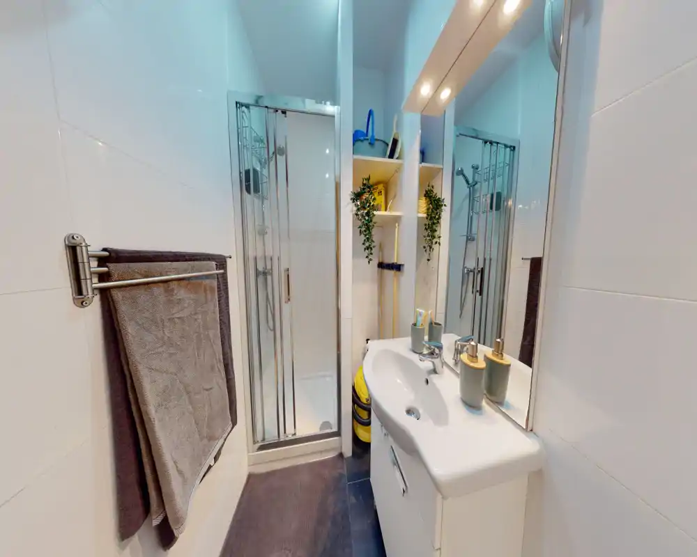 Salle de bain moderne et compacte avec douche vitrée, meuble lavabo blanc, miroir et accessoires rangés — lumineuse et propre.