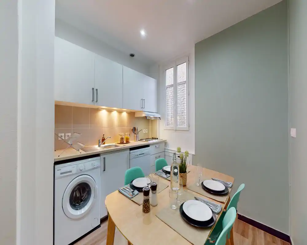 Cuisine moderne et compacte avec électroménagers intégrés et table dressée pour quatre personnes ; l'éclairage sous les placards et une large fenêtre offrent une atmosphère claire et chaleureuse.