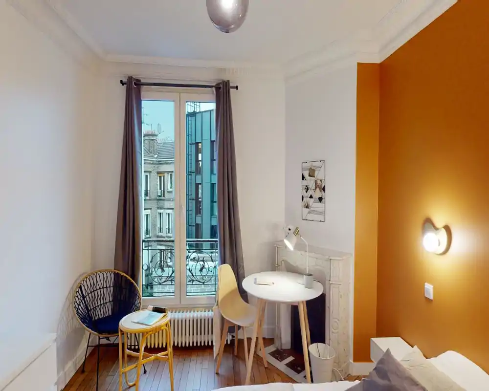 Coin chambre lumineux et chaleureux avec une petite table ronde, une chaise et une lampe de lecture près de la fenêtre. Le mur orange et les assises confortables donnent une ambiance accueillante pour se détendre ou travailler.