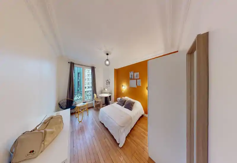 Chambre lumineuse et soignée avec un lit double confortable contre un mur orange chaleureux, parquet, un petit bureau près de la fenêtre et des objets décoratifs — idéale pour un séjour cosy.