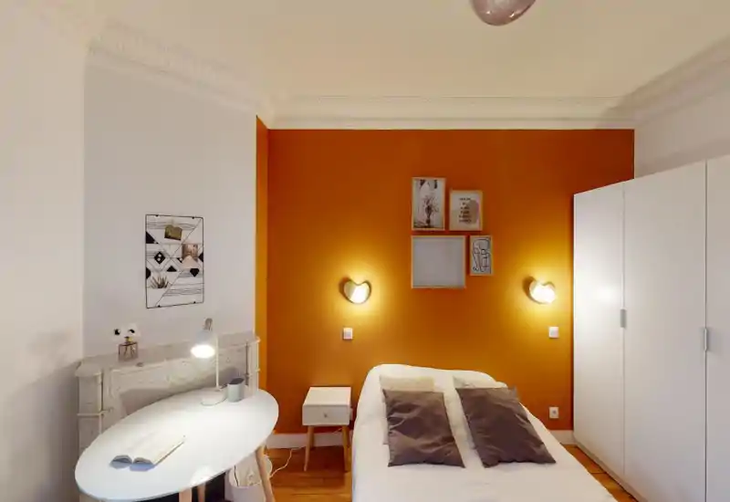 Chambre lumineuse et chaleureuse avec un mur orange, lit double avec coussins, table de chevet et armoire — propre, bien décorée et bien éclairée.