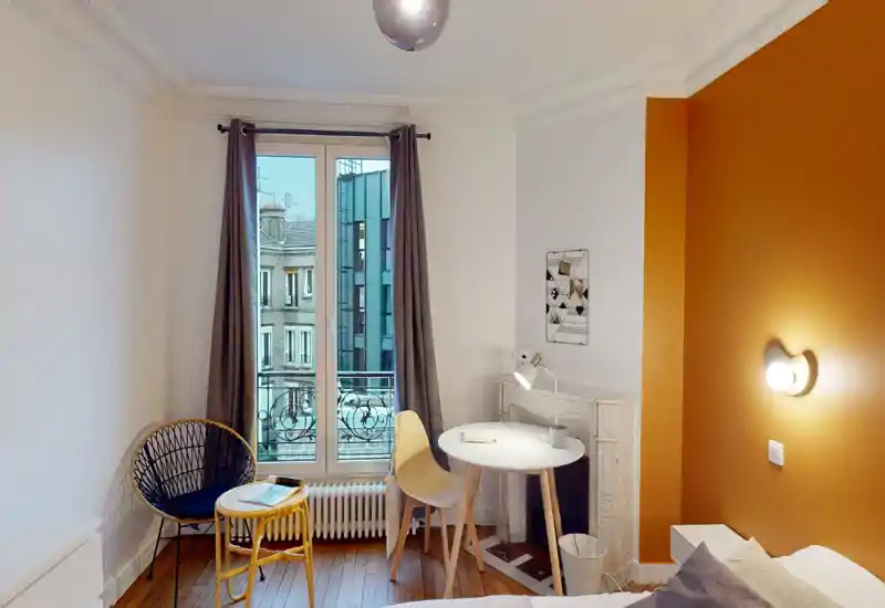 Coin chambre lumineux et chaleureux avec une petite table ronde, une chaise et une lampe de lecture près de la fenêtre. Le mur orange et les assises confortables donnent une ambiance accueillante pour se détendre ou travailler.