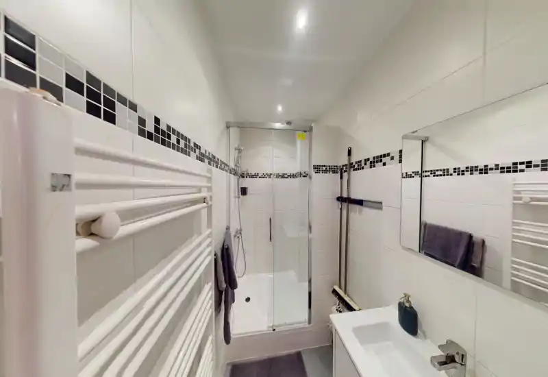 Bright, modern bathroom with a glass-enclosed shower, white tiled walls with black mosaic border, wall-mounted towel radiator and a compact sink with mirror.