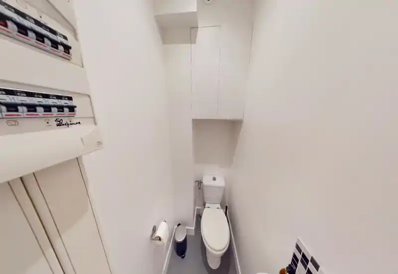 Compact toilet room with a standard cistern, wall-mounted storage above and a small waste bin; clean white walls and tiled floor make it suitable for listings.