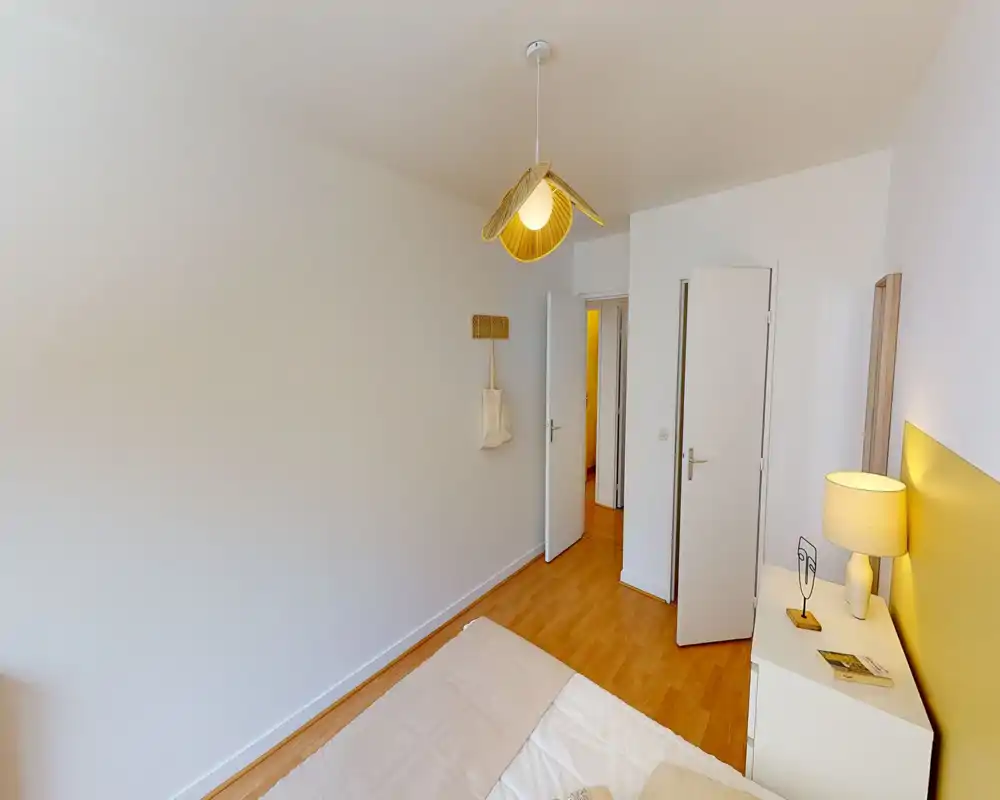 Chambre lumineuse au style épuré avec lit double visible, parquet et table de chevet avec lampe ; murs neutres et éclairage chaleureux pour une ambiance calme.