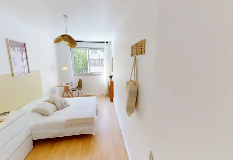 Bright, simply styled bedroom with a double bed, bedside drawers and a small table by the window that provides natural light. The room features warm wooden flooring, neutral linens and minimalist decor for a calm atmosphere.