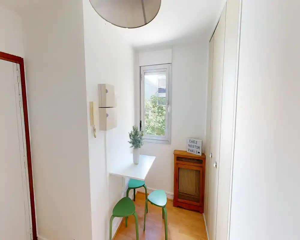 Coin repas compact avec une petite table blanche, trois tabourets verts, une plante décorative et une fenêtre apportant de la lumière naturelle — idéal pour des repas décontractés.