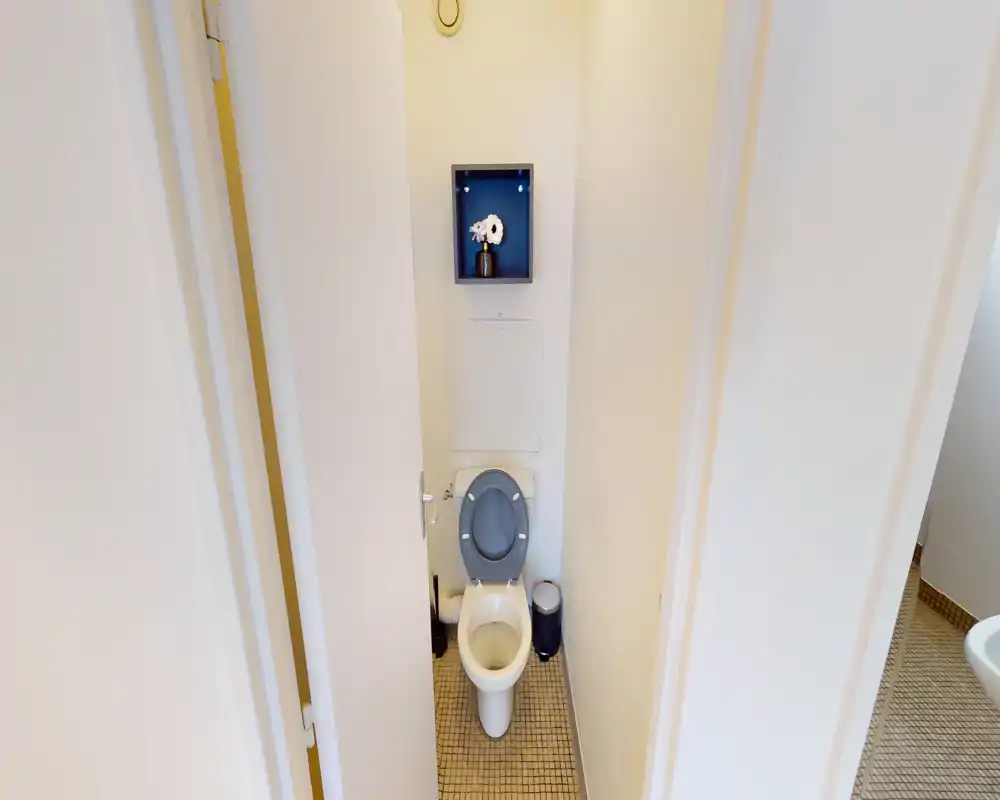 Petite toilette propre avec WC blanc, abattant gris, petite étagère murale et sol carrelé — lumineux et soigné.