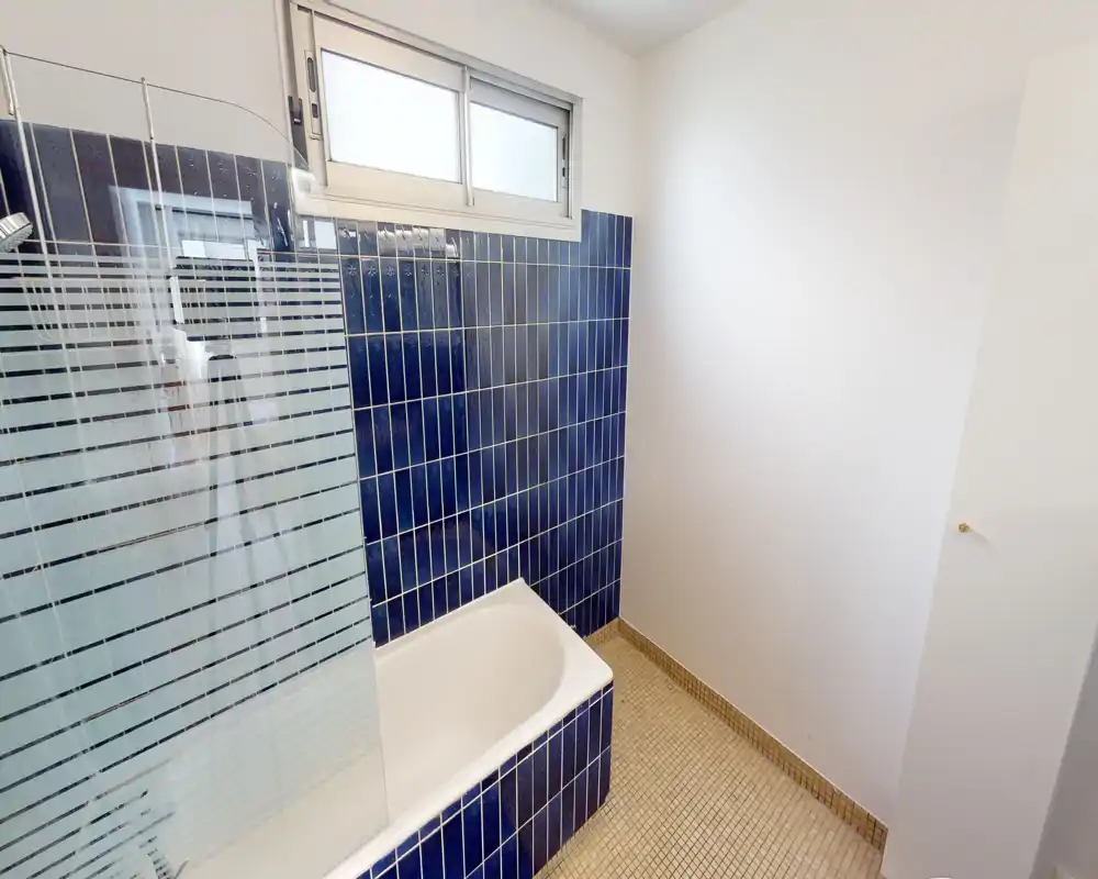 Salle de bain compacte et lumineuse avec baignoire, carreaux céramiques bleus et paroi de douche en verre dépoli sous une fenêtre haute.