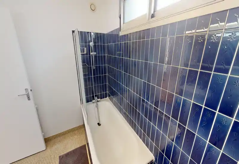 Clean bathroom with a white bathtub and decorative deep-blue wall tiles, lit by a high window. Compact layout with a shower head over the tub.