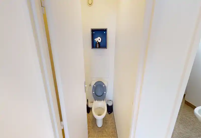 Compact, clean toilet room with a white toilet, gray seat, small wall shelf and tiled floor — bright and tidy.