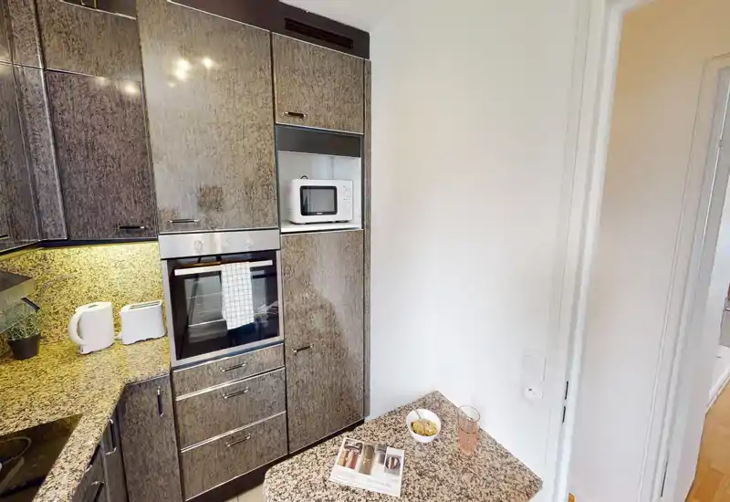 Compact modern kitchen with built-in oven, microwave and granite countertops; small breakfast corner with a magazine and glass, perfect for quick meals.