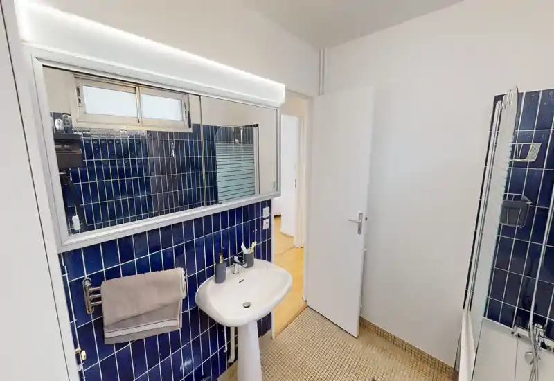 Clean, compact bathroom with a pedestal sink, large mirror, blue tiled wall and a bathtub partially visible on the right. Bright, functional space with a towel rail and toiletries.