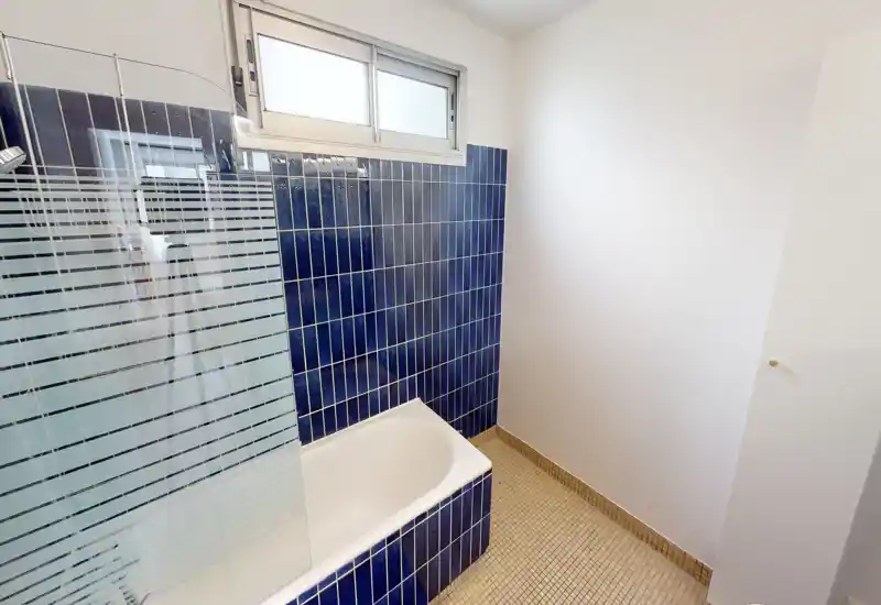 Bright, compact bathroom with a bathtub, blue ceramic tiles and a frosted glass shower screen under a high window.