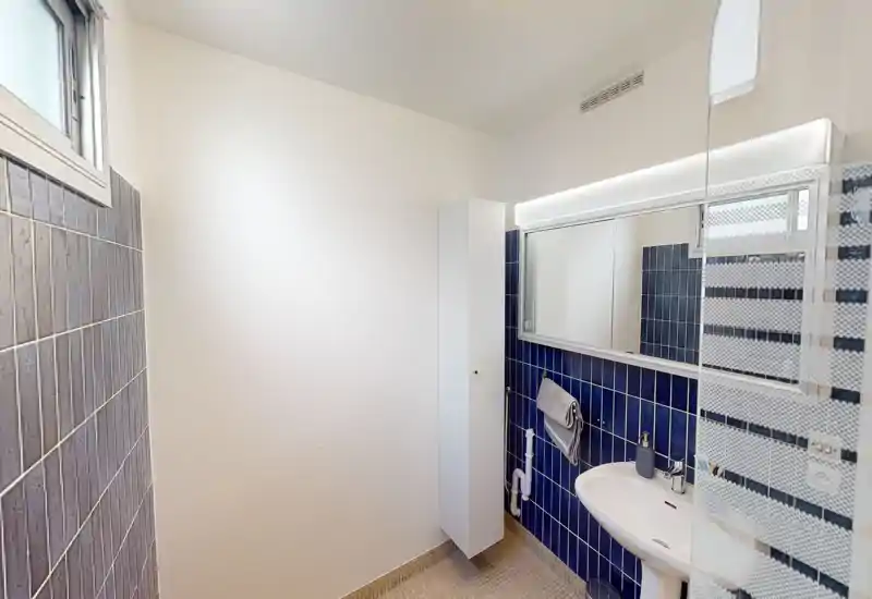 Compact modern bathroom with blue tiled half-wall, wall-mounted sink, mirror cabinet and vertical storage unit. Bright, clean and efficiently arranged.