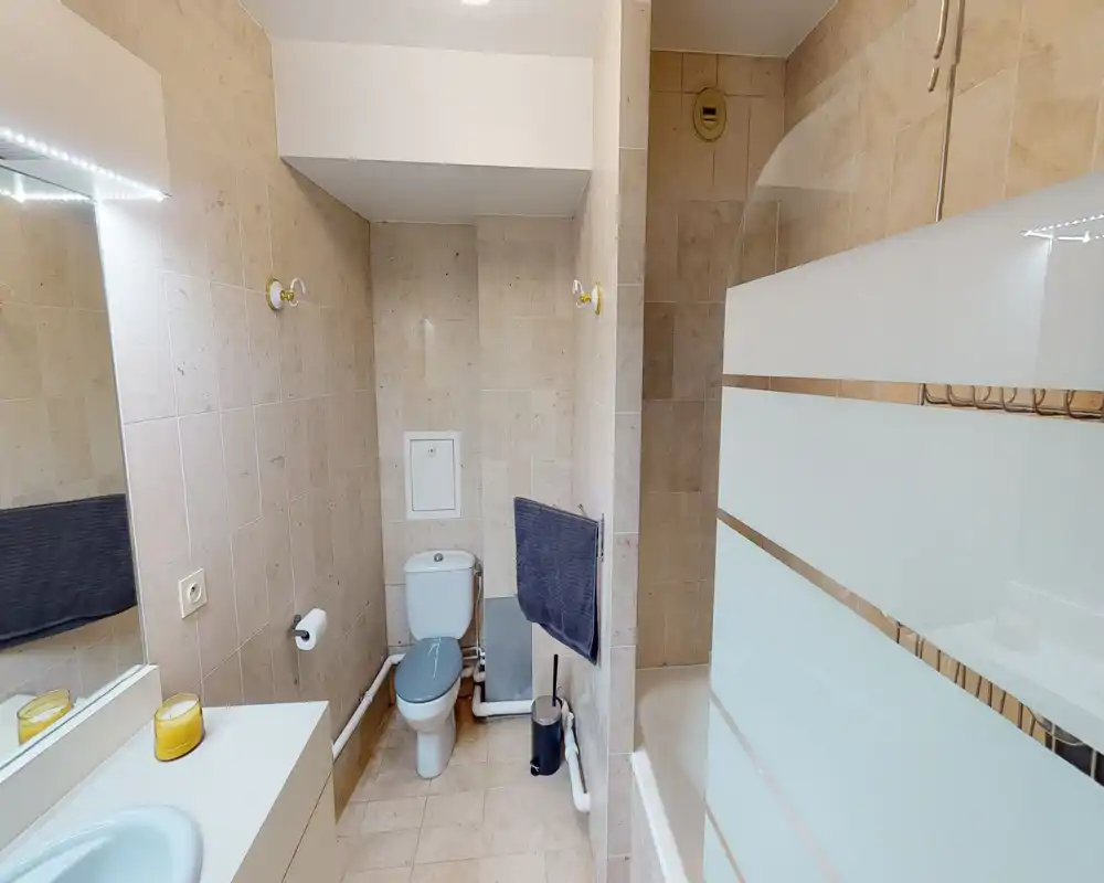 Bagno piastrellato pulito con WC, vasca con separazione in vetro satinato, lavabo e specchio — ben illuminato e ordinato.