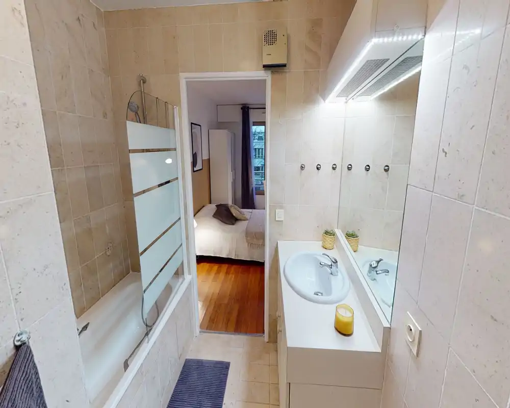 Bagno piastrellato e pulito con vasca, lavabo e grande specchio; ben illuminato e fotografato dalla porta con vista sulla camera da letto adiacente.