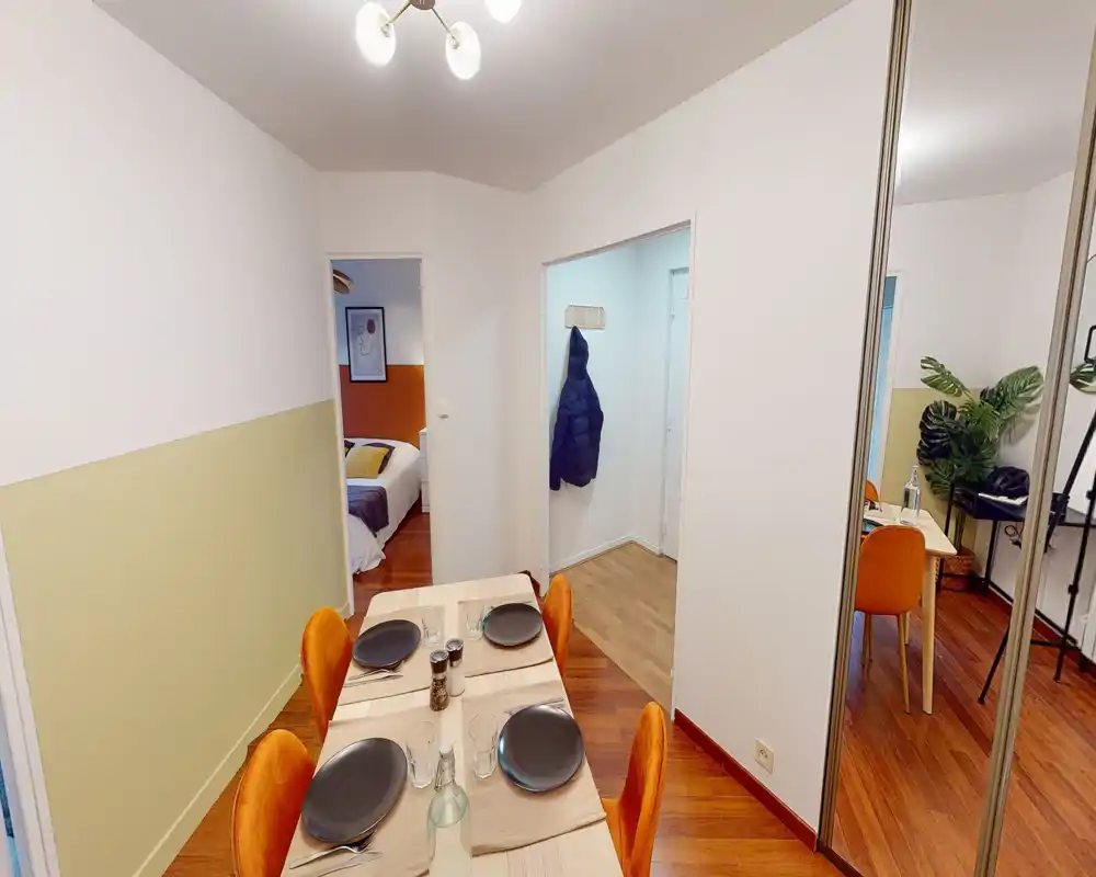 Zona pranzo luminosa e ben arredata con tavolo in legno per quattro e sedie arancioni, adiacente a una camera da letto e al corridoio.