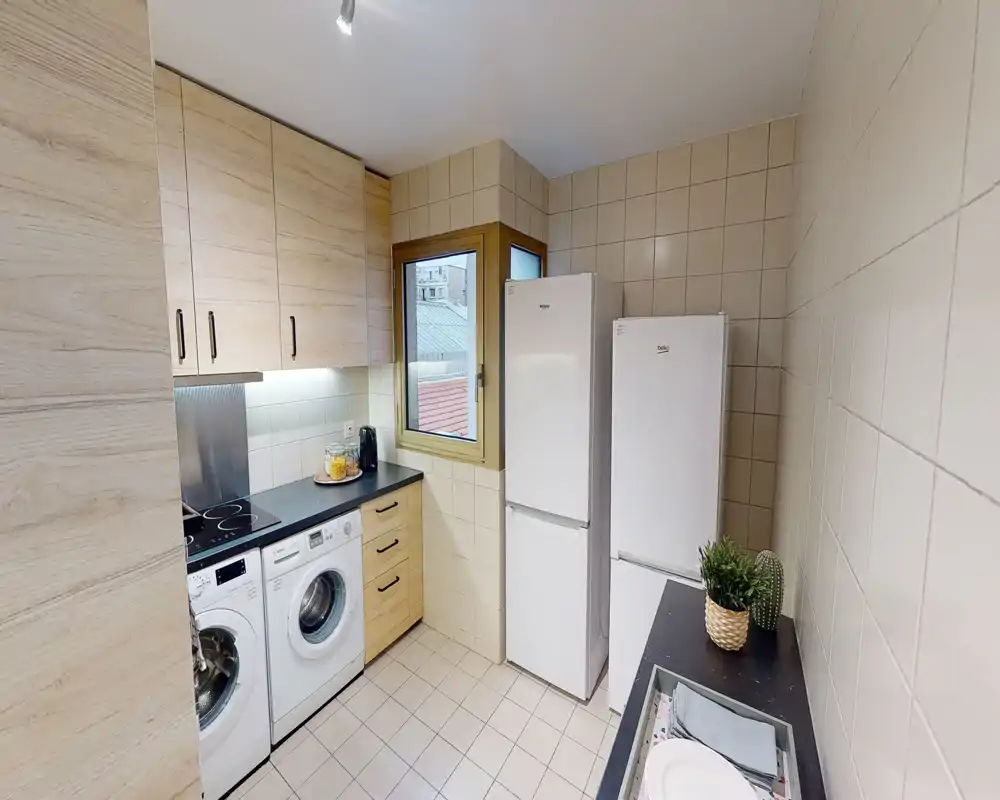Cucina compatta e ben tenuta con elettrodomestici integrati, inclusi lavatrice, piano cottura e due frigoriferi a colonna. Pensili in legno chiaro, pareti piastrellate e una piccola finestra rendono lo spazio luminoso e funzionale, ideale per appartamenti cittadini.